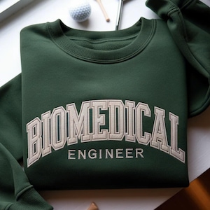 Op de afbeelding: Een donkergroene sweatshirt met de geborduurde tekst "BIOMEDICAL ENGINEER" in lichtbeige, gebogen over de borst. Een golfbal en een tee zijn zichtbaar op de achtergrond.