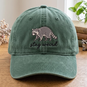 Puede incluir: Una gorra de béisbol verde bosque con un gráfico bordado de un mapache gris y negro caminando. Debajo del mapache, el texto "stay weird" está bordado en una tipografía cursiva negra. La gorra tiene una corona estructurada y visera curva.