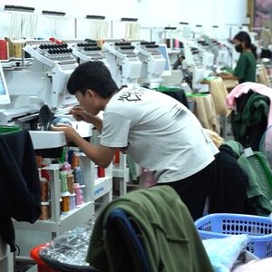 May include: A row of white industrial embroidery machines with multiple spools of colorful thread. Each machine features a digital screen and a needle assembly for stitching designs onto fabric, arranged in a long, well-lit production workshop.
