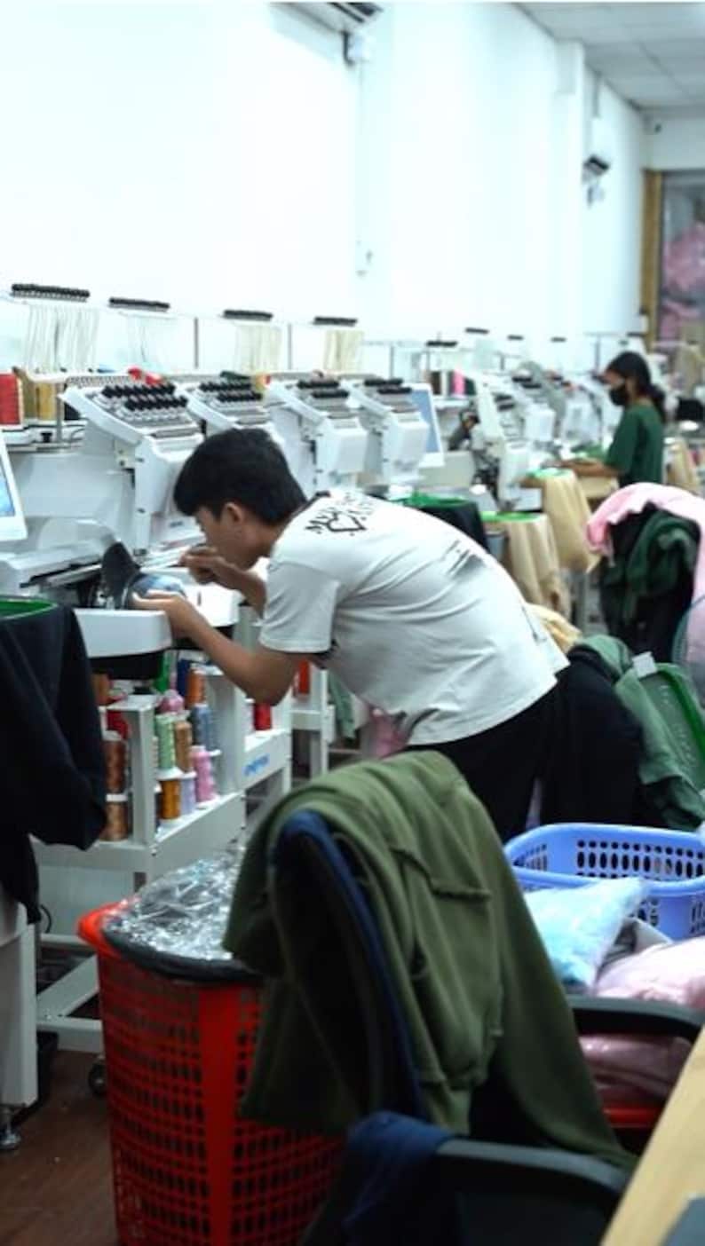 May include: A row of white industrial embroidery machines with multiple spools of colorful thread. Each machine features a digital screen and a needle assembly for stitching designs onto fabric, arranged in a long, well-lit production workshop.