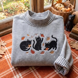 Peut inclure: Un sweat-shirt gris clair doté d'un col montant à motif vichy noir et blanc. L'avant présente une broderie de trois chats noirs jouant avec des feuilles d'automne orange et marron, avec des poignets assortis au motif vichy.