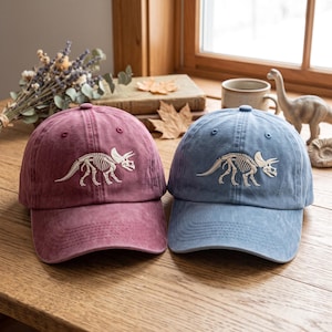 Pode incluir: Dois bonés de beisebol, um em bordô suave e outro em azul poeirento, cada um com um design bordado em branco de um esqueleto de triceratops no painel frontal. Ambos os bonés possuem uma construção de seis painéis e uma aba curva.