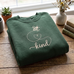 Peut inclure: Un sweat-shirt à col rond vert forêt foncé avec une broderie en fil clair. Le motif représente une petite abeille volant le long d'une ligne en pointillés formant un cœur au-dessus du mot kind écrit en minuscules avec une police cursive.