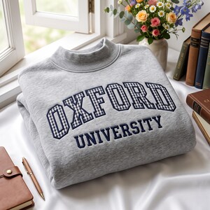 Op de afbeelding: Een lichtgrijs sweatshirt met een opstaande kraag en de tekst OXFORD UNIVERSITY geborduurd op de voorzijde. Het woord OXFORD is uitgevoerd in een marineblauw en wit vichy-ruitpatroon, terwijl UNIVERSITY in effen marineblauwe letters staat.