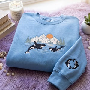 May include: A light blue crewneck sweatshirt featuring an embroidered landscape scene on the chest with two orcas swimming in water, snow-capped mountains, green pine trees, and an orange sun. A smaller embroidery of two orcas appears on the sleeve.