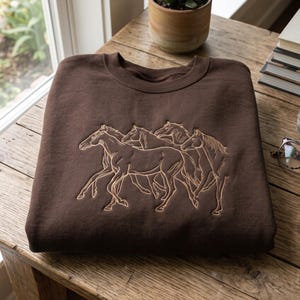 May include: A dark brown crewneck sweatshirt featuring a light tan embroidered line art design of a group of four horses running. The horses are depicted in profile and three-quarter views, showcasing their manes and muscular forms in detail.