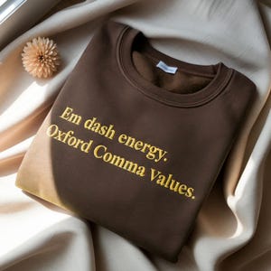 Può includere: Una felpa marrone con la scritta "Em dash energy. Oxford Comma Values." in lettere gialle. La felpa è piegata su un tessuto color crema, con un fiore decorativo nell'angolo in alto a sinistra.