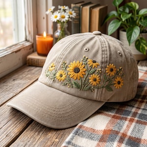 Op de afbeelding: Een beige honkbalpet met gedetailleerd borduurwerk van gele zonnebloemen met groene stengels en bladeren op de voorpanelen. De pet heeft een gebogen klep, gestikte oogjes voor ventilatie en een knoopje boven op de kruin.