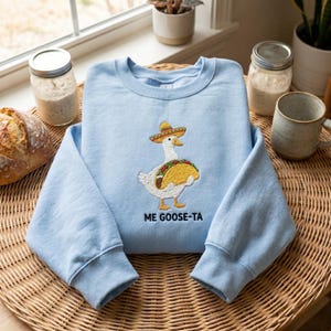 May include: A light blue sweatshirt with an embroidered design of a goose wearing a sombrero and holding a taco. The text "ME GOOSE-TA" is below the design. The sweatshirt is displayed on a woven surface.