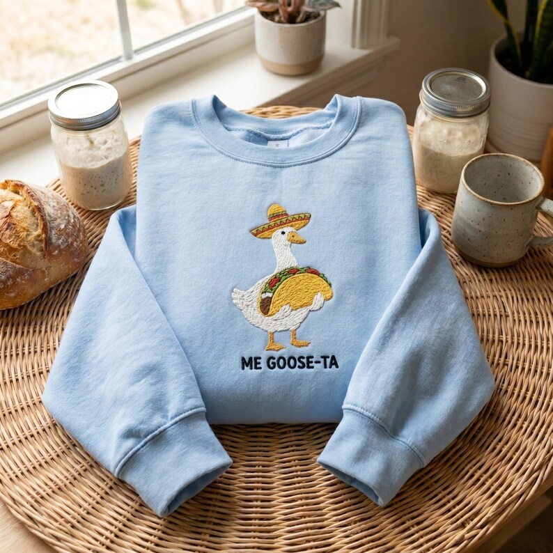 May include: A light blue sweatshirt with an embroidered design of a goose wearing a sombrero and holding a taco. The text "ME GOOSE-TA" is below the design. The sweatshirt is displayed on a woven surface.