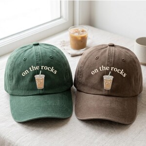May include: Two baseball caps, one dark green and one brown, each featuring an embroidered graphic of an iced coffee in a plastic cup with the text "on the rocks" arched above the illustration in a white, rounded, sans-serif font.