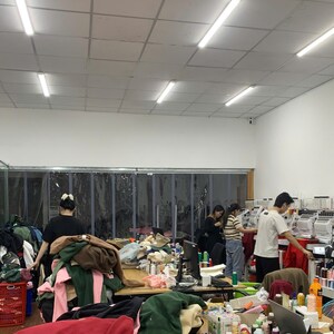May include: A workspace featuring multiple embroidery machines, a large wooden table covered in piles of colorful fabric garments, and several individuals working in a bright room with white walls and a drop ceiling with fluorescent lighting.