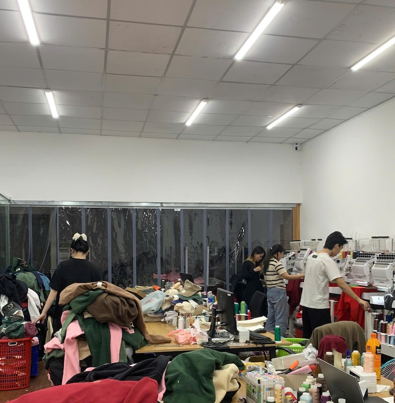 May include: A workspace featuring multiple embroidery machines, a large wooden table covered in piles of colorful fabric garments, and several individuals working in a bright room with white walls and a drop ceiling with fluorescent lighting.