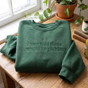 Könnte beinhalten: Ein waldgrüner Rundhals-Pullover mit dem gestickten Text "i was told there would be pickles" in passendem Dunkelgrün. Das Kleidungsstück hat lange Ärmel mit gerippten Bündchen sowie einen gerippten Halsausschnitt und zeigt ein schlichtes Design.