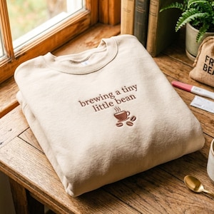 Puede incluir: Una sudadera de cuello redondo en color beige con bordado marrón en el pecho. El texto "brewing a tiny little bean" aparece sobre un gráfico bordado de una taza de café humeante y tres granos de café centrados debajo.