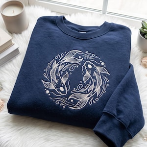 Könnte beinhalten: Ein marineblaues Langarm-Sweatshirt mit einer kreisförmigen Stickerei, die zwei Koi-Fische in einer Yin-Yang-Formation zeigt. Die Stickerei ist in hellem Cremefaden gehalten und betont die Schuppen, Flossen und die wirbelnden Wassermotive.