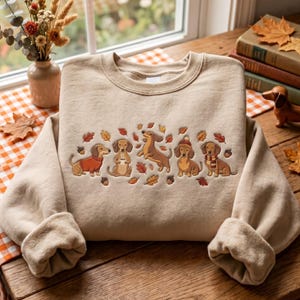May include: A beige crewneck sweatshirt featuring an embroidered design of five dachshunds in various poses, including one wearing a sweater, one holding a mug, and one wearing a beanie, surrounded by scattered autumn leaves and small brown acorns.