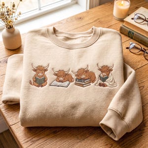 May include: A beige crewneck sweatshirt featuring an embroidered design of four brown Highland cows in various poses with books. One cow wears glasses and reads, one lies on an open book, one sleeps on a stack of books, and one sits wrapped in a blanket.