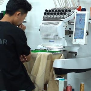 May include: A white industrial embroidery machine with multiple thread spools and a digital screen displaying a graphic design. The machine is positioned over a beige fabric garment, with various colored thread spools stored below.