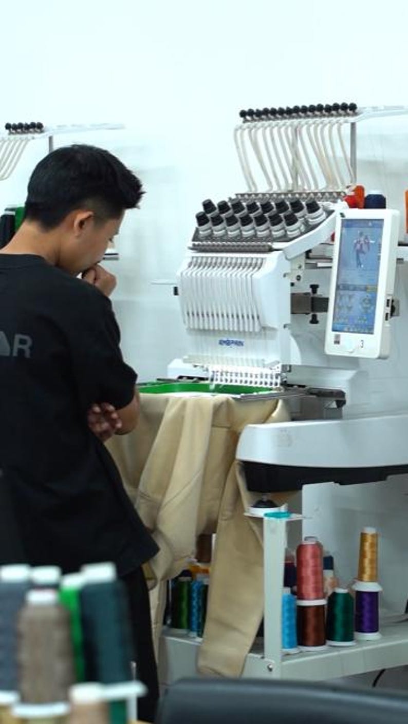 May include: A white industrial embroidery machine with multiple thread spools and a digital screen displaying a graphic design. The machine is positioned over a beige fabric garment, with various colored thread spools stored below.