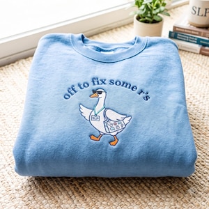 May include: A light blue crewneck sweatshirt featuring an embroidered graphic of a white goose wearing sunglasses, a teal lanyard with an SLP badge, and carrying a bag with medical icons. The text above the goose reads, off to fix some r's.