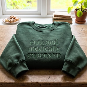 Peut inclure: Un sweat-shirt à col rond vert forêt avec le texte brodé "cute and medically expensive" dans un fil vert foncé assorti. Le vêtement présente des manches longues, une encolure côtelée et une coupe classique avec une finition soignée.