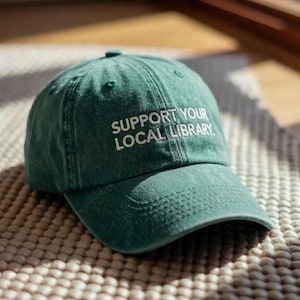 Peut inclure: Une casquette de baseball vert forêt avec une finition délavée de style vintage. L'avant de la casquette présente le texte blanc brodé "SUPPORT YOUR LOCAL LIBRARY". Le chapeau a une couronne structurée, une visière incurvée et un bouton.