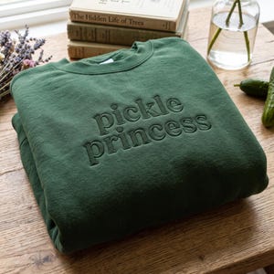 Peut inclure: Un sweat-shirt à col rond vert forêt avec le texte brodé pickle princess en fil vert foncé assorti. Le vêtement présente un col côtelé et des manches longues, présenté plié sur une surface en bois.