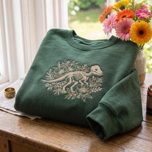 Op de afbeelding: Een bosgroene sweater met ronde hals en een crèmekleurig borduursel van een dinosaurusskelet omringd door bloemmotieven. Het borduursel is gecentreerd op de borst van het kledingstuk, dat lange mouwen en een geribbelde halslijn heeft.
