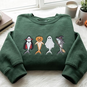 Könnte beinhalten: Ein dunkelgrüner Rundhals-Pullover mit einer Stickerei von vier Figuren: ein Hai als Vampir verkleidet, ein Hai mit Kürbiskopf, ein weißes Gespenst und ein Hai mit Hexenhut, der einen Besenstiel in der Flosse hält.