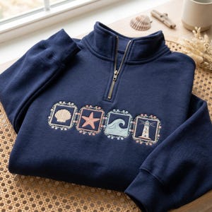 Puede incluir: Sudadera azul marino con cremallera de un cuarto de largo con diseños costeros bordados: una concha, una estrella de mar, una ola y un faro. La sudadera tiene una cremallera dorada y está hecha de un material suave. El diseño está enmarcado por bordados florales.