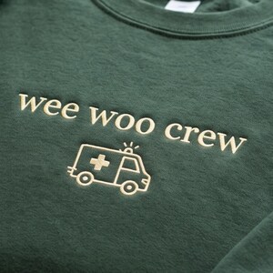 May include: A dark forest green crewneck sweatshirt featuring the embroidered text "wee woo crew" in a light cream color. Below the text is a simple line-art embroidery of an ambulance with a cross symbol on the side, also in light cream.