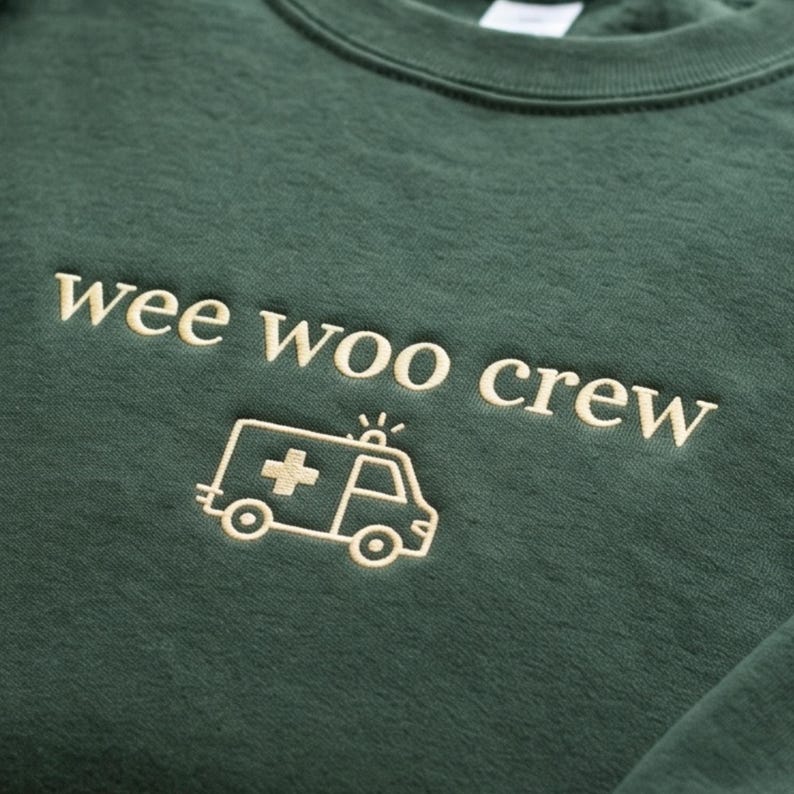May include: A dark forest green crewneck sweatshirt featuring the embroidered text "wee woo crew" in a light cream color. Below the text is a simple line-art embroidery of an ambulance with a cross symbol on the side, also in light cream.