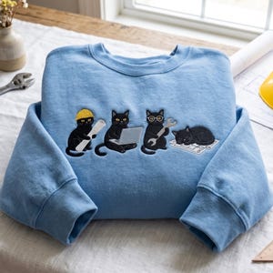 May include: A light blue crewneck sweatshirt featuring an embroidered graphic of four black cats. One cat wears a yellow hard hat and holds a blueprint, another uses a laptop, a third wears glasses and holds a spanner, and the fourth sleeps on a plan.