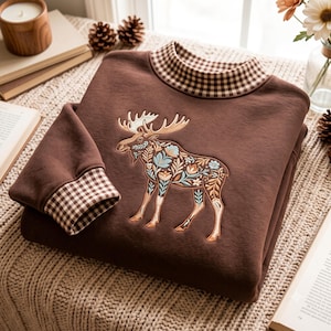 Könnte beinhalten: Ein braunes Sweatshirt mit hohem Kragen und Ärmelbündchen in braun-weißem Vichy-Karo. In der Mitte des Kleidungsstücks befindet sich eine Stickerei eines Elches, der mit detaillierten Blumen- und Blattmotiven in Blau- und Beigetönen gefüllt ist.