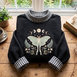 Op de afbeelding: Een zwart sweatshirt met lange mouwen, een hoge kraag en boorden met een zwart-wit gingham patroon. Op het midden van de borst staat een borduursel van een lichtgroene nachtvlinder, omringd door botanische elementen en maanfasen.