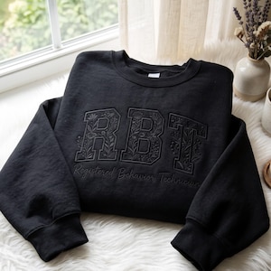 May include: A black crewneck sweatshirt featuring large, block-style letters RBT embroidered on the front. The letters are filled with a detailed floral pattern. Below the letters, the text Registered Behavior Technician is embroidered in script.