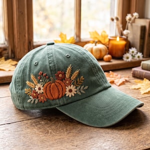 May include: A sage green baseball cap featuring detailed embroidery of a central orange pumpkin surrounded by wheat stalks, small white daisies, and rust-colored flowers. The cap has a curved brim and a structured crown with stitched eyelets.