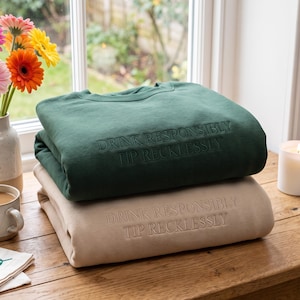 Puede incluir: Una sudadera verde oscuro apilada sobre una sudadera beige. Ambas prendas presentan el texto bordado DRINK RESPONSIBLY TIP RECKLESSLY centrado en el pecho. Las sudaderas tienen cuello redondo y mangas largas, dobladas cuidadosamente.