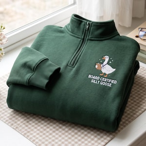 May include: A dark green quarter-zip sweatshirt featuring an embroidered graphic of a white goose wearing glasses and a lanyard while holding a clipboard. Below the goose, the text reads BOARD CERTIFIED SILLY GOOSE in white letters.