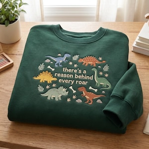 May include: A dark green crewneck sweatshirt featuring an embroidered design of various dinosaurs, including a blue, yellow, green, and brown dinosaur, surrounding the text "there's a reason behind every roar" with small bone and leaf accents.