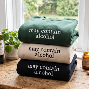 Op de afbeelding: Een stapel van drie sweaters met ronde hals in donkergroen, beige en zwart. Elke sweater is op de borst bedrukt met de tekst "may contain alcohol" in een eenvoudig, strak lettertype, waarbij de tekstkleur contrasteert met de stof.
