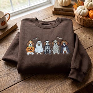 Könnte beinhalten: Ein dunkelbraunes Sweatshirt mit Rundhalsausschnitt und einer Stickerei von fünf Hunden in Halloween-Kostümen, darunter ein Kürbis, ein Geist, ein Skelett, eine Mumie und ein Vampir, begleitet von zwei kleinen weißen Fledermäusen darüber.