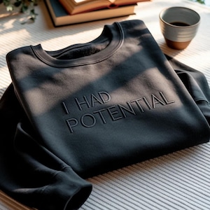 May include: Dark gray sweatshirt with the embroidered text "I HAD POTENTIAL". The crew neck sweatshirt is folded on a gray surface, with a book and a cup of coffee in the background. The sweatshirt has ribbed cuffs and a relaxed fit.
