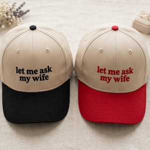 May include: Two baseball caps with the text "let me ask my wife". One cap has a black brim and a black button, the other has a red brim and a red button. The caps are tan with the text in black and red.