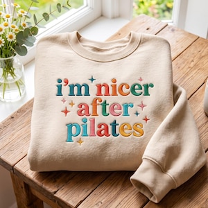 Puede incluir: Una sudadera de cuello redondo en color beige con el texto bordado "i'm nicer after pilates" en una fuente multicolor. El diseño incluye pequeños detalles de estrellas bordadas en varios colores alrededor del texto en la parte frontal.