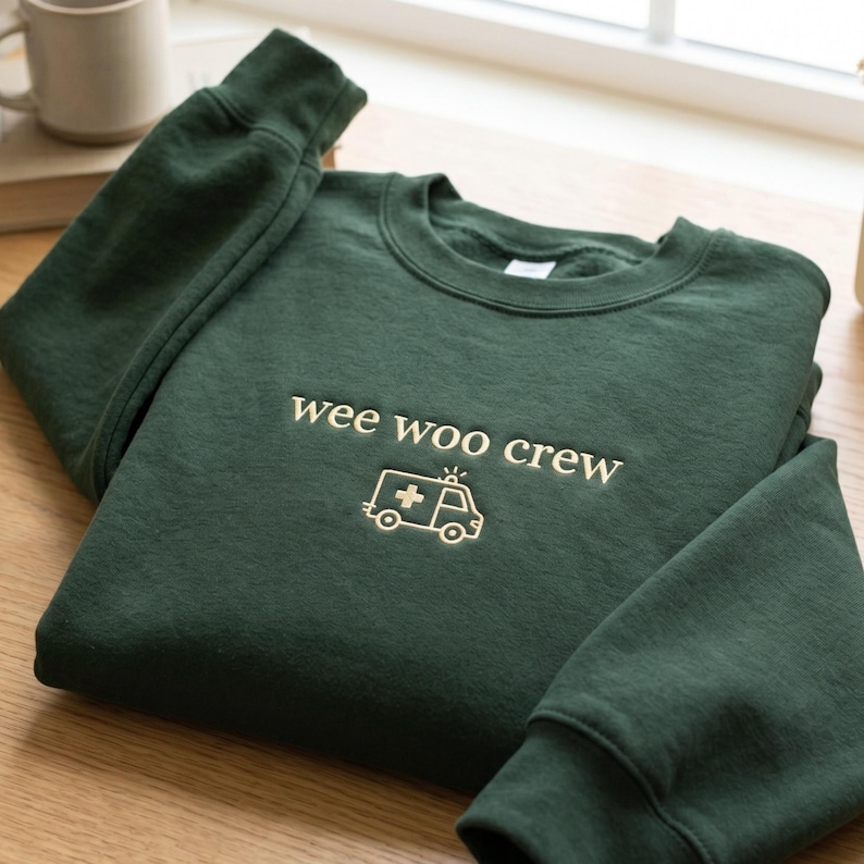 May include: A dark forest green crewneck sweatshirt featuring the embroidered text "wee woo crew" in a light cream color, positioned above a simple line-art graphic of an ambulance with a cross symbol on its side.