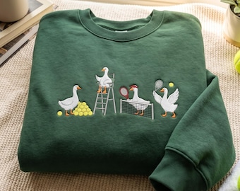 Puede incluir: Una sudadera de cuello redondo verde oscuro con un bordado de cuatro gansos blancos jugando al tenis. Un ganso está sentado en una silla de árbitro, otro junto a pelotas amarillas, uno golpea una pelota sobre una red y otro sostiene una raqueta.