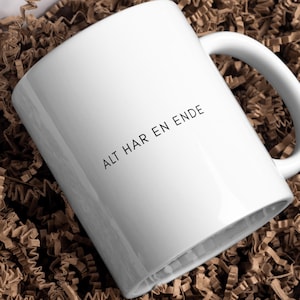 May include: White ceramic mug with a curved handle. The mug features the text "ALT HAR EN ENDE" in black, centered on the side. The mug is set against a brown, textured background.