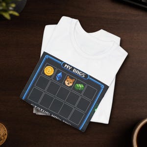 Peut inclure: Un t-shirt blanc avec un graphique en pixel art représentant un écran d'inventaire numérique intitulé MY BAGS. L'écran affiche des icônes pour Bitcoin, Ethereum, un chien Shiba Inu et une liasse de billets, avec le texte STATUS: HODLING en bas.